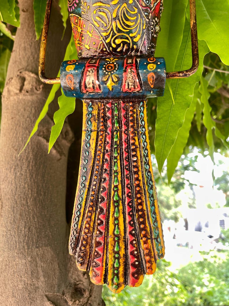 May include: A colorful wooden bird ornament with intricate details and a hanging loop. The bird is painted in shades of blue, yellow, red, green, and brown, with a variety of patterns and designs.