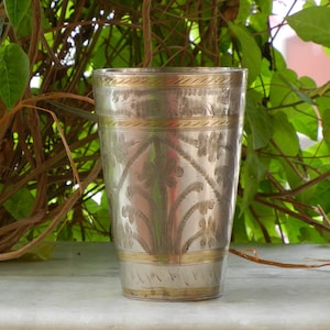 May include: A silver drinking glass with an ornate design. The glass has a gold band around the top and bottom. The design is etched into the metal and features floral motifs.