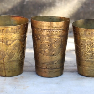 May include: Three gold-colored metal drinking glasses with intricate etched designs. The glasses are all the same size and shape, and they have a traditional, ornate look.