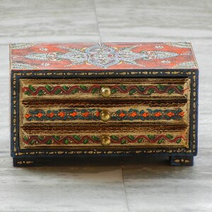 Puede incluir: Una caja de madera con tres cajones, pintada con intrincados dise&ntilde;os florales en tonos rojos, naranjas, verdes y dorados. La caja tiene un borde azul oscuro y tiradores de color dorado.