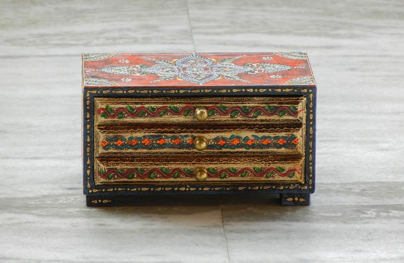 Puede incluir: Una caja de madera con tres cajones, pintada con intrincados dise&ntilde;os florales en tonos rojos, naranjas, verdes y dorados. La caja tiene un borde azul oscuro y tiradores de color dorado.