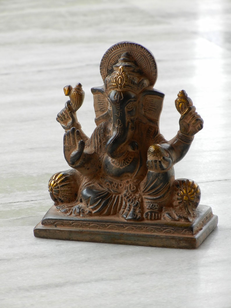 Puede incluir: Una estatua de Ganesha negra y dorada con una base decorativa. La estatua est&aacute; sentada sobre una superficie de m&aacute;rmol blanco.