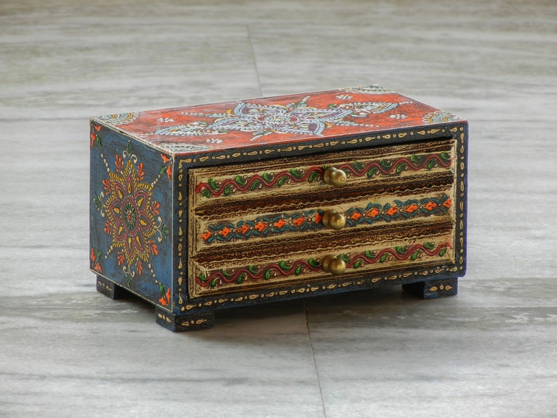 Puede incluir: Una peque&ntilde;a caja de madera con tres cajones pintados con intrincados dise&ntilde;os florales en tonos azules, naranjas, rojos y dorados. La caja tiene pomos de lat&oacute;n en cada caj&oacute;n.