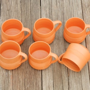 May include: Six orange ceramic mugs with a textured surface. The mugs have handles and are arranged in a semi-circle on a wooden surface.