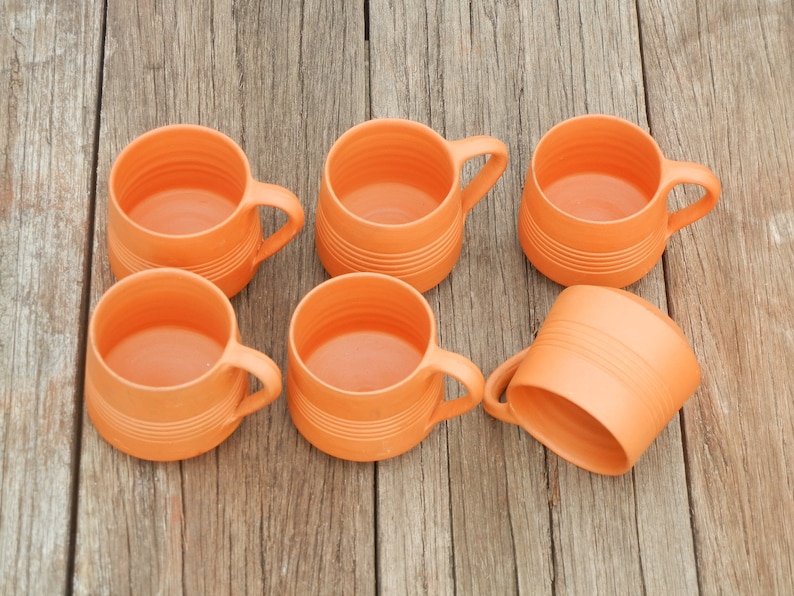 May include: Six orange ceramic mugs with a textured surface. The mugs have handles and are arranged in a semi-circle on a wooden surface.