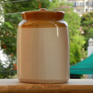 May include: A white ceramic jar with a brown lid. The jar has a smooth, glossy finish. The lid is slightly domed and has a textured surface. The jar is sitting on a wooden surface. Jaipur Treasures is printed on the surface.