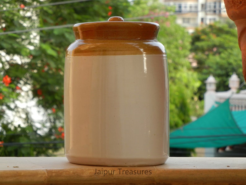 May include: A white ceramic jar with a brown lid. The jar has a smooth, glossy finish. The lid is slightly domed and has a textured surface. The jar is sitting on a wooden surface. Jaipur Treasures is printed on the surface.