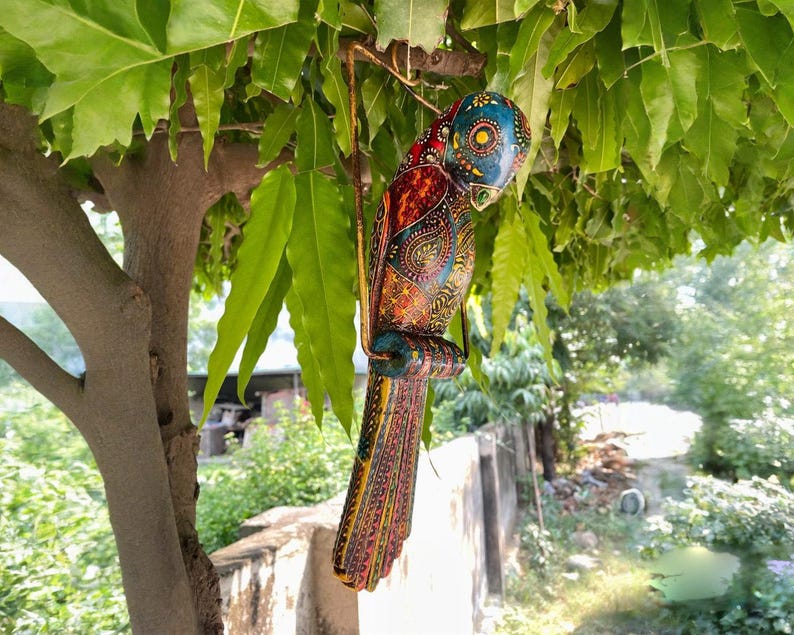 May include: A colorful wooden parrot with intricate painted details, hanging from a branch. The parrot is painted in shades of blue, green, red, yellow, and orange.