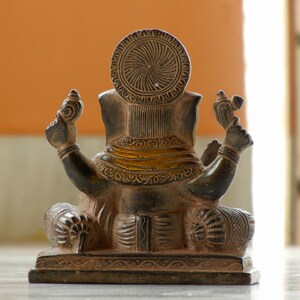Puede incluir: Una estatua de Ganesha, el dios hind&uacute; de la sabidur&iacute;a y la prosperidad, de color marr&oacute;n oscuro tallada de forma intrincada, sentada en una postura meditativa. La estatua est&aacute; hecha de metal y tiene un ribete dorado alrededor de la cintura.