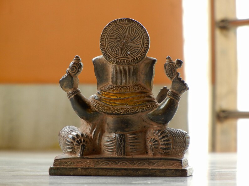Puede incluir: Una estatua de Ganesha, el dios hind&uacute; de la sabidur&iacute;a y la prosperidad, de color marr&oacute;n oscuro tallada de forma intrincada, sentada en una postura meditativa. La estatua est&aacute; hecha de metal y tiene un ribete dorado alrededor de la cintura.
