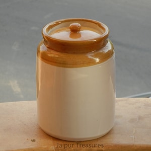 May include: A white ceramic jar with a brown lid. The jar has a brown band around the top. The jar is sitting on a wooden surface. Jaipur Treasures is printed on the surface.