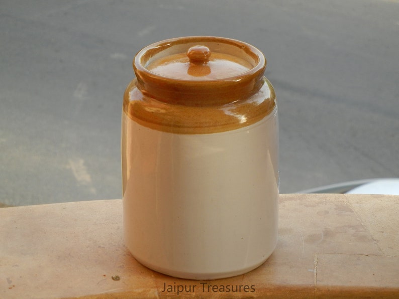 May include: A white ceramic jar with a brown lid. The jar has a brown band around the top. The jar is sitting on a wooden surface. Jaipur Treasures is printed on the surface.