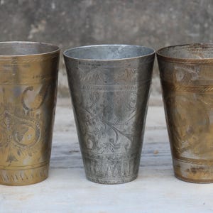 May include: Three ornate metal drinking cups with intricate designs. The cups are made of brass and silver and have a traditional Indian style.