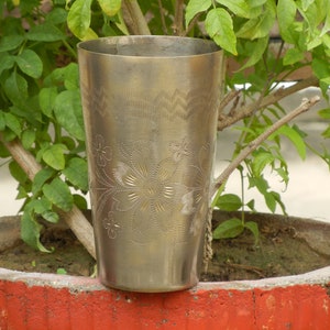 May include: A silver metal drinking glass with a floral design etched into the surface. The glass is sitting on a red planter.