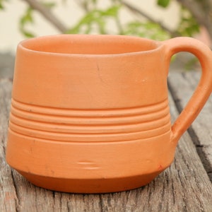 May include: A terracotta mug with a rounded handle and a textured, striped design. The mug is sitting on a wooden surface.