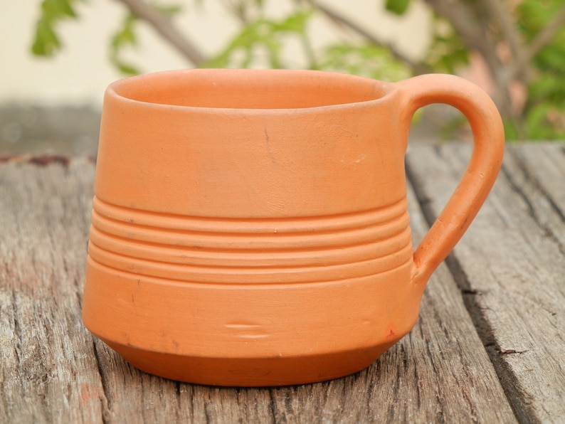 May include: A terracotta mug with a rounded handle and a textured, striped design. The mug is sitting on a wooden surface.