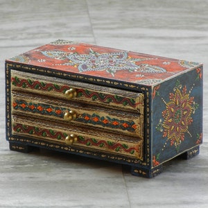 Puede incluir: Una peque&ntilde;a caja de madera con tres cajones, pintada con intrincados dise&ntilde;os florales en tonos de rojo, naranja, azul, verde y dorado. La caja tiene una base azul oscuro y detalles dorados.