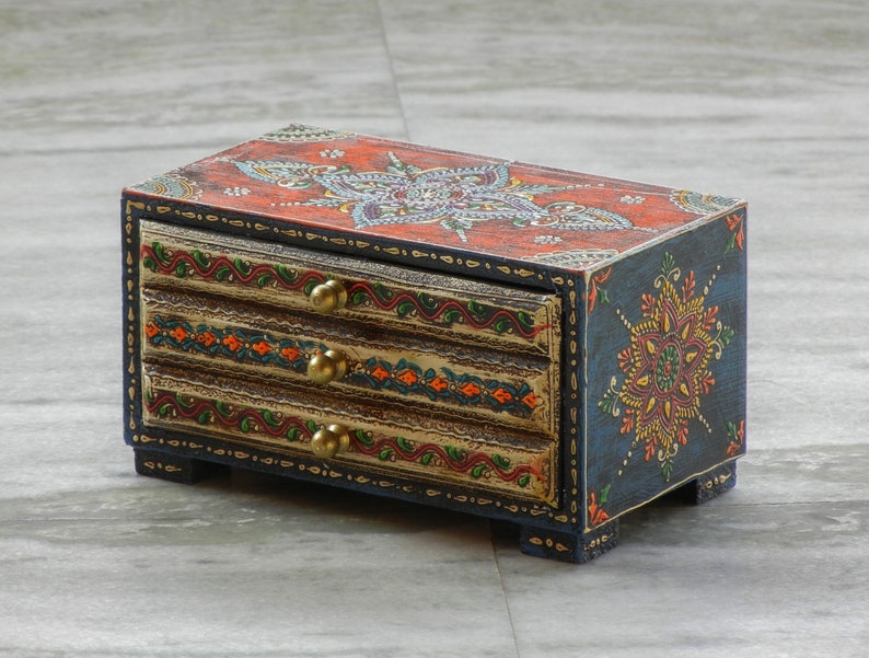Puede incluir: Una peque&ntilde;a caja de madera con tres cajones, pintada con intrincados dise&ntilde;os florales en tonos de rojo, naranja, azul, verde y dorado. La caja tiene una base azul oscuro y detalles dorados.