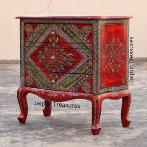May include: A red wooden chest of drawers with three drawers. The drawers are decorated with intricate floral patterns in shades of green, gold, and purple. The chest is accented with gold trim and sits on four ornate legs.