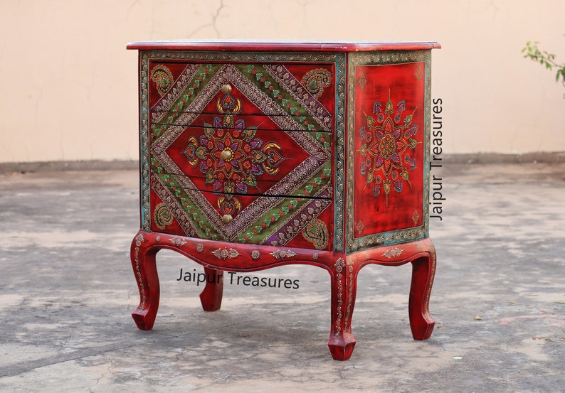 May include: A red wooden chest of drawers with three drawers. The drawers are decorated with intricate floral patterns in shades of green, gold, and purple. The chest is accented with gold trim and sits on four ornate legs.