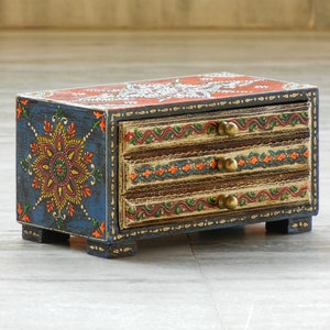 Puede incluir: Una peque&ntilde;a caja de madera con tres cajones pintados con intrincados dise&ntilde;os florales en tonos azules, naranjas, amarillos y verdes. Los cajones tienen pomos de lat&oacute;n y est&aacute;n decorados con un patr&oacute;n floral.