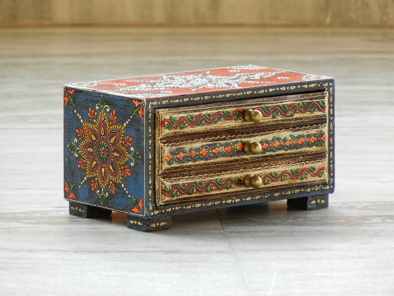 Puede incluir: Una peque&ntilde;a caja de madera con tres cajones pintados con intrincados dise&ntilde;os florales en tonos azules, naranjas, amarillos y verdes. Los cajones tienen pomos de lat&oacute;n y est&aacute;n decorados con un patr&oacute;n floral.