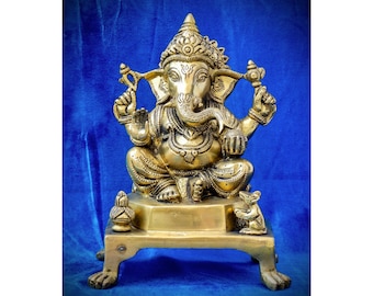 May include: A gold-colored brass statue of Ganesha, the Hindu god of beginnings, sitting on a square base with a rat at his feet. The statue is on a blue velvet background.