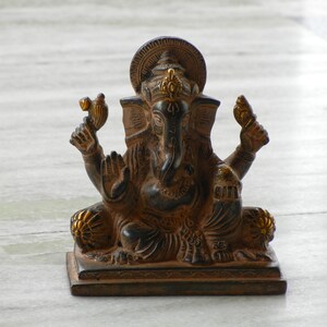 Puede incluir: Una estatua de Ganesha negra y dorada con una base decorativa. La estatua est&aacute; sentada sobre una superficie de m&aacute;rmol blanco.