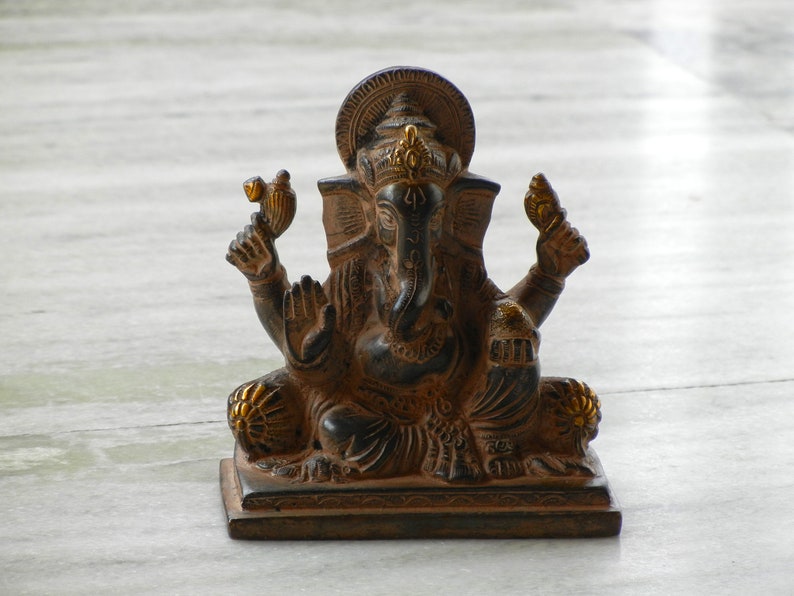 Puede incluir: Una estatua de Ganesha negra y dorada con una base decorativa. La estatua est&aacute; sentada sobre una superficie de m&aacute;rmol blanco.