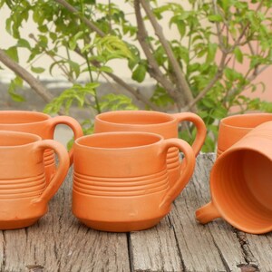 May include: Six terracotta mugs with handles, arranged on a wooden surface. The mugs have a rustic, earthy appearance and are perfect for serving beverages.