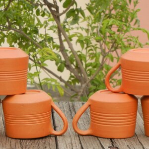 May include: Six terracotta mugs stacked in two groups of three. The mugs are a warm orange color and have a textured, ribbed surface.