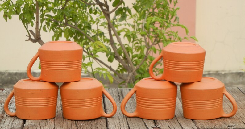 May include: Six terracotta mugs stacked in two groups of three. The mugs are a warm orange color and have a textured, ribbed surface.