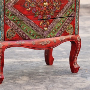 May include: A red wooden chest of drawers with intricate hand-painted details in gold, green, and purple. The chest has two drawers and sits on four ornate legs.