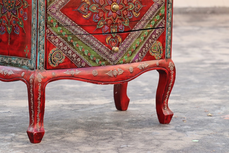 May include: A red wooden chest of drawers with intricate hand-painted details in gold, green, and purple. The chest has two drawers and sits on four ornate legs.