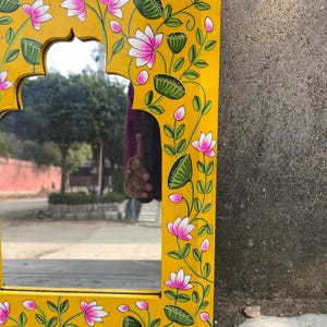 Puede incluir: Un espejo de madera amarillo con un dise&ntilde;o floral pintado a mano. El espejo tiene una forma de arco tradicional y est&aacute; decorado con flores rosas y hojas verdes.