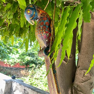 May include: A colorful wooden parrot figurine with intricate details and a bright blue, red, yellow, and green color scheme. The parrot is hanging from a branch with a wire.