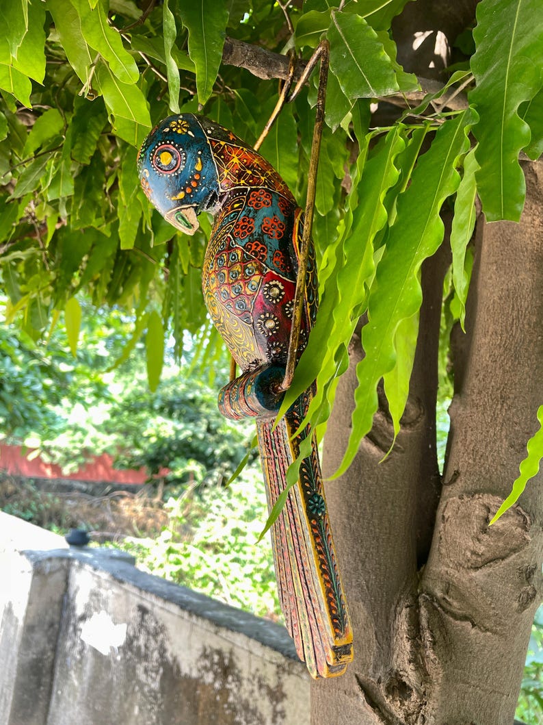 May include: A colorful wooden parrot figurine with intricate details and a bright blue, red, yellow, and green color scheme. The parrot is hanging from a branch with a wire.