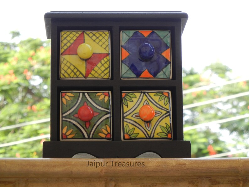 May include: A black wooden box with four small drawers. Each drawer has a colorful ceramic front with a different geometric pattern and a knob. The box is sitting on a wooden surface. Jaipur Treasures.