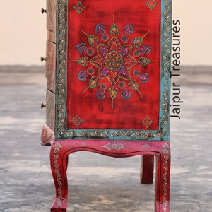 May include: A red wooden chest of drawers with intricate hand-painted floral designs in shades of blue, green, and gold. The chest has four drawers and sits on four ornate legs. The text "Jaipur Treasures" is visible on the side of the chest.
