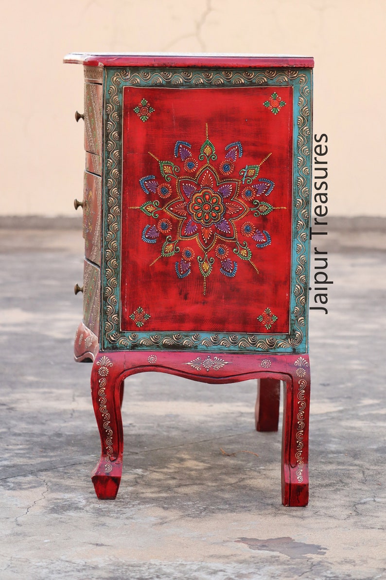 May include: A red wooden chest of drawers with intricate hand-painted floral designs in shades of blue, green, and gold. The chest has four drawers and sits on four ornate legs. The text "Jaipur Treasures" is visible on the side of the chest.