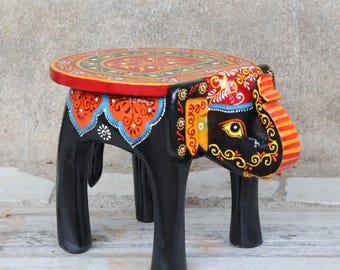 May include: A hand-painted wooden elephant stool. The stool features a round top with a red border and intricate patterns in orange, green, and yellow. The elephant-shaped base is black with colorful details, including an orange and blue design.