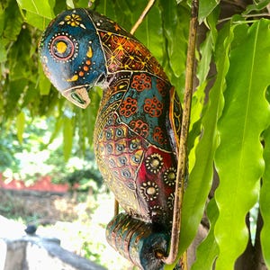 May include: A colorful wooden parrot ornament with intricate patterns and details. The parrot is painted in shades of blue, green, red, yellow, and gold. It is hanging from a metal hook.