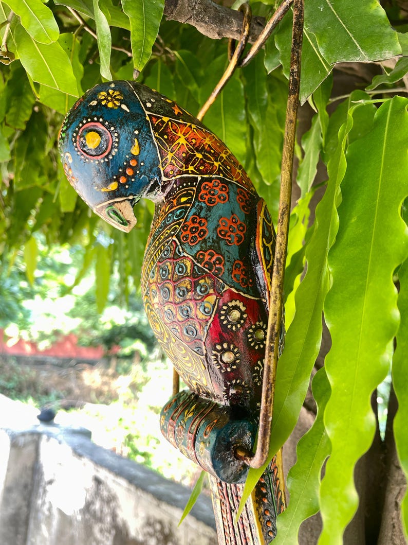 May include: A colorful wooden parrot ornament with intricate patterns and details. The parrot is painted in shades of blue, green, red, yellow, and gold. It is hanging from a metal hook.