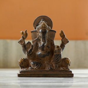 Puede incluir: Una estatua de Ganesha negra y dorada con un dise&ntilde;o detallado. La estatua est&aacute; sentada sobre una base y tiene un halo decorativo.