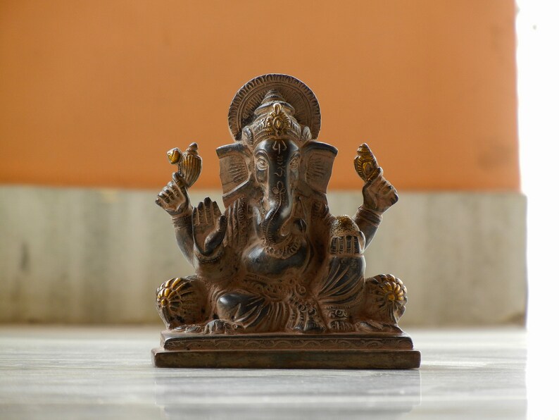 Puede incluir: Una estatua de Ganesha negra y dorada con un dise&ntilde;o detallado. La estatua est&aacute; sentada sobre una base y tiene un halo decorativo.