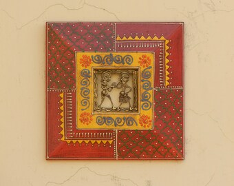 Könnte beinhalten: Quadratische, handbemalte Holz-Wandkunst mit einem zentralen Metallrelief, das zwei Figuren darstellt. Der Rahmen ist rot mit gemusterten Abschnitten in Rot, Gelb und Grün. Das Kunstwerk ist quadratisch.