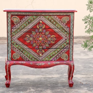May include: A red wooden chest of drawers with three drawers. The drawers are decorated with intricate floral patterns in shades of green, gold, and purple. The chest has ornate legs and a carved top.