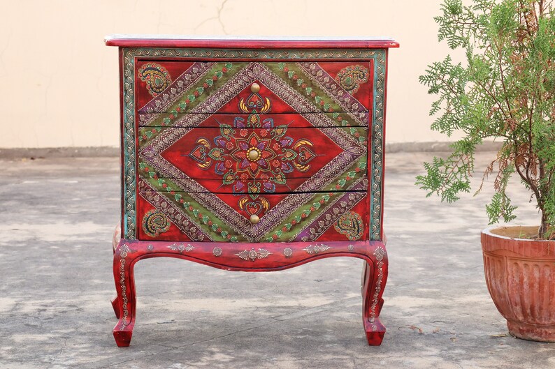 May include: A red wooden chest of drawers with three drawers. The drawers are decorated with intricate floral patterns in shades of green, gold, and purple. The chest has ornate legs and a carved top.