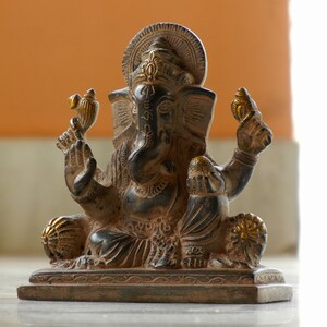 Puede incluir: Una estatua de Ganesha de color marr&oacute;n oscuro y dorado con un dise&ntilde;o detallado. La estatua est&aacute; sentada sobre una base con un borde decorativo.
