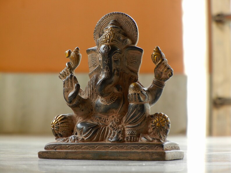 Puede incluir: Una estatua de Ganesha de color marr&oacute;n oscuro y dorado con un dise&ntilde;o detallado. La estatua est&aacute; sentada sobre una base con un borde decorativo.